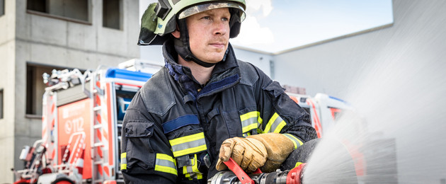 Ein Feuerwehrmann in Feuerwehrschutzkleidung hält ein Strahlrohr in der Hand und versprüht Wasser. Im Hintergrund steht ein Feuerwehrfahrzeug.