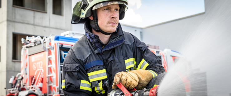 Ein Feuerwehrmann in Feuerwehrschutzkleidung hält ein Strahlrohr in der Hand und versprüht Wasser. Im Hintergrund steht ein Feuerwehrfahrzeug.