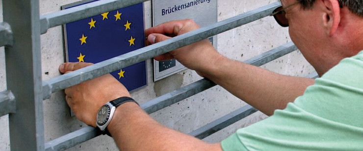 Mitarbeiter befestigt viereckiges Schild der EU-Flagge an Brückengeländer
