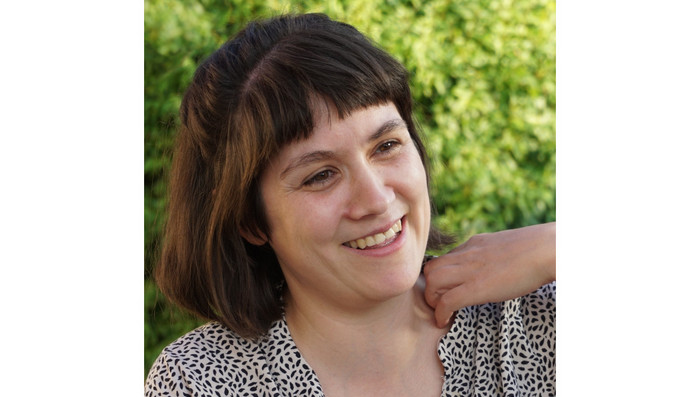 Porträt von Christiane Haas. Eine Frau mit schulterlangen, braunen Haaren blickt lächelnd zur Seite und berührt mit einer Hand ihre linke Schulter.