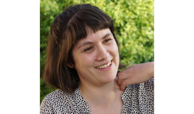 Porträt von Christiane Haas. Eine Frau mit schulterlangen, braunen Haaren blickt lächelnd zur Seite und berührt mit einer Hand ihre linke Schulter.