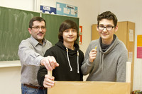 Zwei Schüler stehen mit einem Lehrer hinter Ihrem Forschungsprojekt. Ein Junge hält eine Nudel in der Hand.