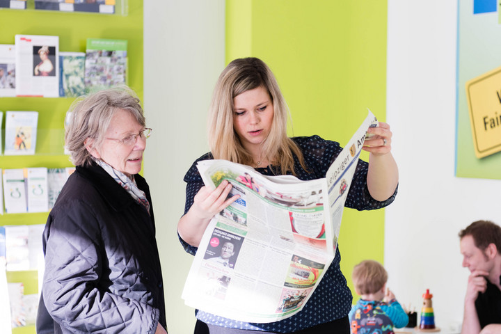Eine jüngere und eine ältere Frau blicken zusammen in eine Zeitung, im Hintergrund spielt ein Vater mit seinem Sohn