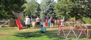 Auf einer Wiese mit Rutsche und einem Klettergerüst stehen Kindergarten- und Krippenkinder mit Erwachsenen in einem Kreis. 