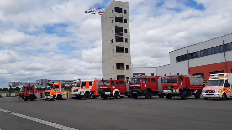 Neun verschiedene Rettungsfahrzeuge stehen nebeneinander aufgereiht vor einem Gebäude mit Turm der Rettungswache