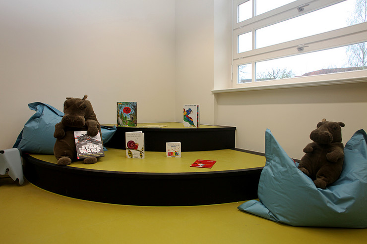 Podest in einer Ecke am Fenster der Kinderbibliothek in Plagwitz mit einer Stufe und grünem Fußboden, darauf zwei Plüschtiere, Kissen und Medien.