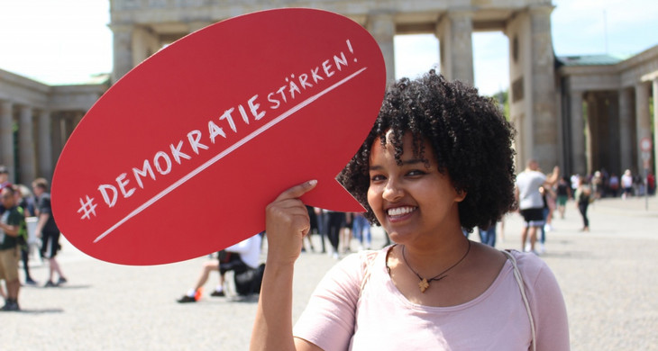 Junge Frau vor dem Brandenburger Tor zu sehen, die eine rote Sprechblase mit der Aufschrift "#Demokratie stärken!" hält.