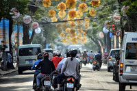Ansicht auf zwei Motorräder von hinten, welche durch eine Straße fahren, die Fahrer tragen keine Helme. Die Straße ist gesäumt von parkenden Autos und großen grünen Bäumen, die mit gelb-orangenen Blumenketten mit Lichtern verbunden sind