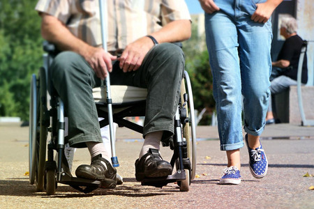 Ein ältere Person sitzt mit einem Gehstock in der Hand im Rollstuhl. Daneben läuft eine andere Person.