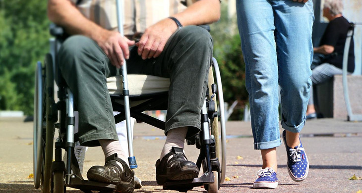 Ein ältere Person sitzt mit einem Gehstock in der Hand im Rollstuhl. Daneben läuft eine andere Person.