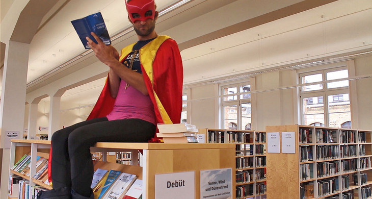 Ein Mann in einem Superheldenkostüm sitzt auf einem Bücherregal in einer Bücherei