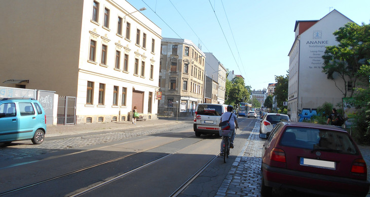 Ansicht der Dieskaustraße mit fahrenden und parkenden Autos und einem Radfahrer. In der Mitte liegen Straßenbahngleise, daneben ist die Straße gepflastert.