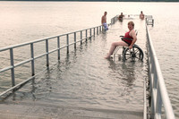 Frau auf Rollstuhl nutzt den Rollstuhlbadesteg um ins Wasser zu gelangen.