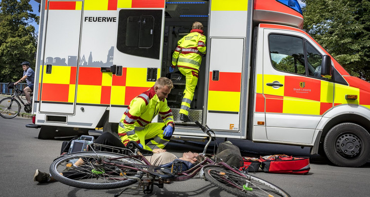 Zwei Notfallsanitäter im Einsatz mit dem Rettungswagen. Eine Frau ist vom Fahrrad gestürzt und liegt am Boden. Die Einsatzkräfte behandeln sie.