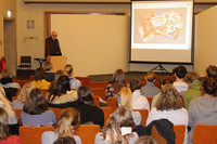 Eine Gruppe Studierender sitzt in mehreren Reihen in einem Saal. Vor ihnen steht ein Mann an einem Pult und zeigt eine Präsentation auf einer Leinwand.