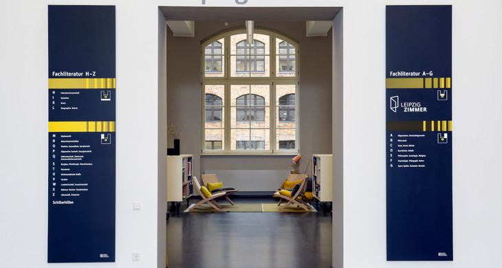 Der Eingang zum LeipzigZimmer ist mit grauer Schrift gekennzeichnet. Man schaut in einen Raum mit Stühlen, Regalen und einem großen Fenster.