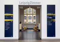 Der Eingang zum LeipzigZimmer ist mit grauer Schrift gekennzeichnet. Man schaut in einen Raum mit Stühlen, Regalen und einem großen Fenster.