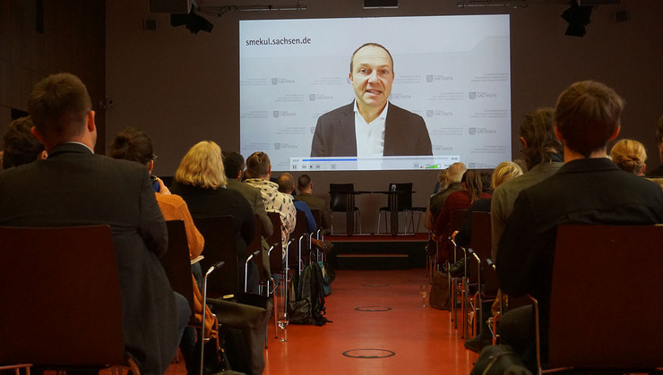 Auf die Bühnenrückseite ist groß ein Video projiziert in dem der sächsische Staatsminister für Landwirtschaft die Teilnehmenden begrüßt