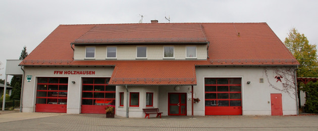 Ein Weißes Haus mit drei großen, roten Rolltoren und einem roten Dach. In den drei Garagen stehen Feuerwehrfahrzeuge.