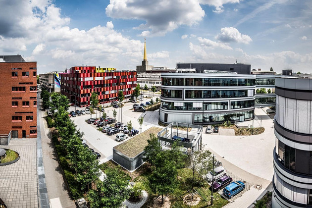 Viele Gebäude auf dem Campus der BioCity in Leipzig. Dazwischen Bäume und parkende Autors und ein Himmel mit einzelnen Wolken.