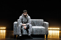 Ein Mann in grauen Sachen auf einem grauem Sofa, dass auf einer Bühne steht.