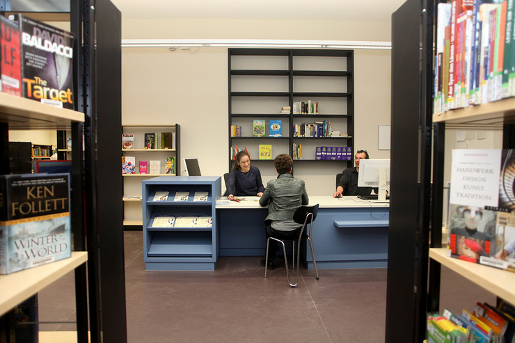 Blick durch die Regale auf einen Beratungsplatz in der Bibliothek Plagwitz mit blauer Theke, dahinter zwei Mitarbeiter, eine Besucherin informiert sich