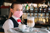 Eine Kellnerin mit Alltagsmaske trägt auf einem Tablett einen Latte Macchiato 