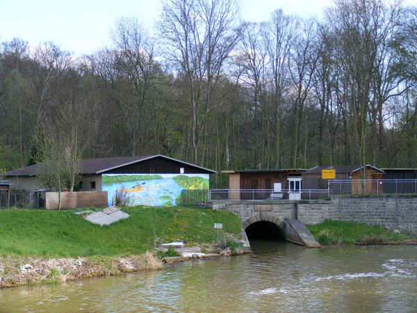 Fluss Pleiße und dahinter einige flache Gebäude der Wassersportanlage Am Pleißewehr