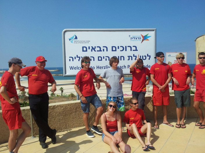 Eine Gruppe junger Menschen mit roten Shirts steht am hellen Sandstrand am Mittelmeer