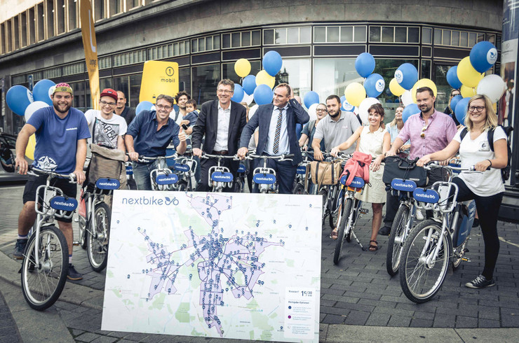Gruppe von Fahrradfahren steht mit Nextbike-Fahrrädern vor einer Karte auf der Verleihstationen und die flexiblen Rückgabezonen von Nextbike markiert sind.