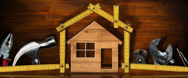 in der Mitte eine Holzschablone in Form eines Hauses, umrandet von einem Zollstock, ebenfalls in Hausform zurechtgebogen. Im Hintergrund diverses Werkzeug wie Hammer und Zangen