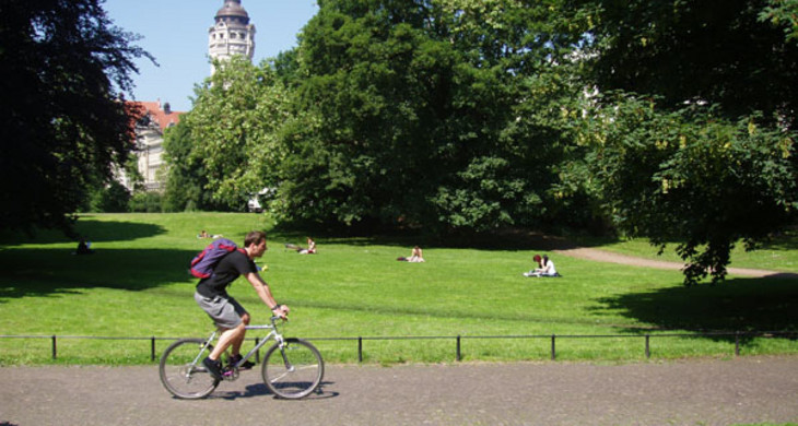 Fahrradfahrer auf einem Weg in einem Leipziger Park. Hinter einer Wiese und Bäumen ist der Turm des Neuen Rathauses sichtbar.