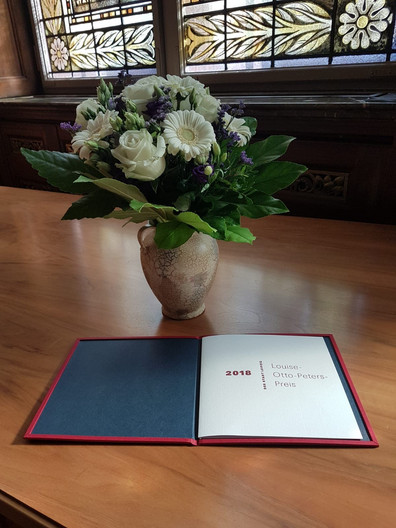 Eine Urkunde mit dem Schriftzug "Louise-Otto-Peters-Preis 2018" und eine Vase mit Blumen auf einem Holztisch