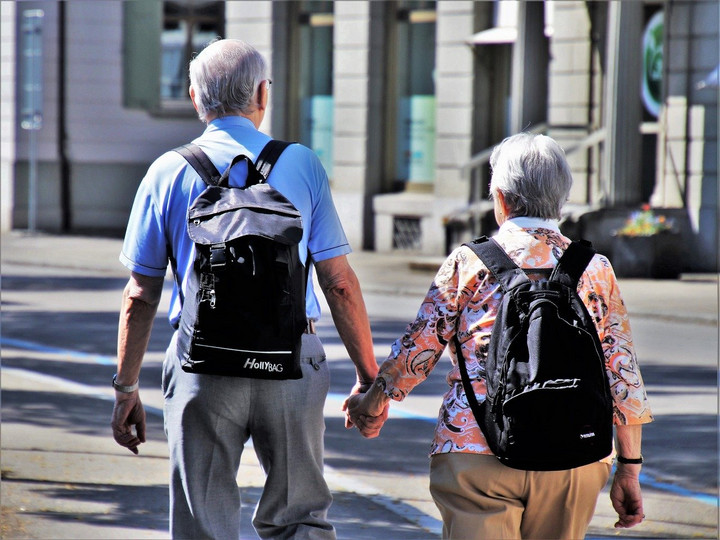 Ein älteres Paar mit grauen Haaren, die sich an der Hand halten. Beide haben schwarze Rucksäcke auf.