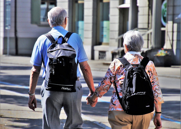 Ein älteres Paar mit grauen Haaren, die sich an der Hand halten. Beide haben schwarze Rucksäcke auf.