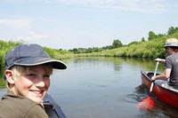 Ein blonder junge am linken Bildrand schaut lachend in die Kamera. Vor ihm befindet sich ein Kanal mit grünem Ufer sowie rechts ein Paddler in einem Boot.