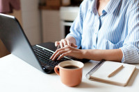 Ein Laptop auf einem Küchentisch, an dem eine Frau arbeitet. Daneben steht eine Tasse Kaffee und liegt ein Notizblock. 