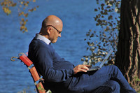 Ein älterer Mann mit Glatze und Brille im blauen Pullover mit einem Laptop auf dem Schoß sitzt auf einem roten Gartenstuhl vor einem See