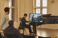 Ein Frau mit Munde-Nasen-Bedeckung spielt Klavier und wird dabei von zwei Männern für einen Livestream gefilmt. 