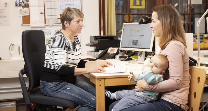 Eine mit dem Baby auf dem Schoß und eine weitere Frau ihr gegenüber sitzen an einem Schreibtisch in einem Büro.
