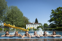 Fünf junge Menschen lehnen in einem Freibad im Pool am Beckenrand und lächeln in die Kamera.