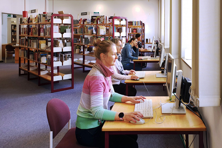 An mehreren Schreibtischn arbeiten Menschen am Computer, im Hintergrund Bücherregale.