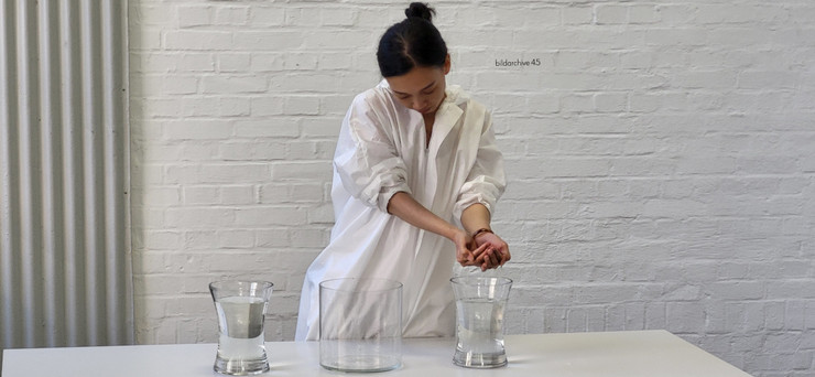 Eine Dame in einem weißen Anzug steht hinter einem weißen Tisch in einem hellen und weißen Raum. Auf dem Tisch stehen drei Glasbehälter mit Wasser. Die Dame entnimmt aus einem Behälter das Wasser.