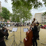 An einer Gedenkstätte spielen vier Musikerinnen vor Zuschauenden, im Hintergrund Häuserfassaden.