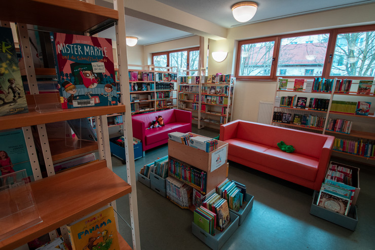 Blick in die Kinderbibliothek mit zwei Couches, Büchertrögen und Regalen. Im Hintergrund mehrere Fenster.