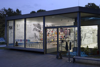 Außenansicht der Galerie für Zeitgenössische Kunst während der blauen Stunde. Durch die großen Glasscheiben sind beleuchtete Räume sichtbar.