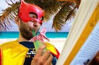 Ein Mann mit roter Superhelden Gesichtsmaske sitzt mit einem Getränk und Buch am Strand vor einer Palme