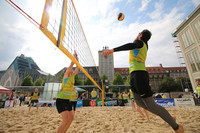 Ein Volleyballspieler springt auf einem Sandplatz zum Ball, um ihn über das Netz zu schlagen, im Hintergrund sieht man Gebäude der Leipziger Innenstadt (Paulinum, Krochhochhaus, ein Teil der Oper)