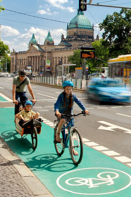 Verkehrsszene in Leipzig vor dem Neuen Rathaus mit Bus, Straßenbahnhaltestelle, Autos, Fahrrädern und Fußgängern. Eine Familie fährt mit ihren Rädern auf einem grünen Radfahrweg.