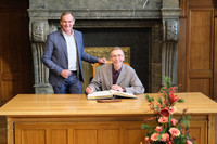 Svante Pääbo sitzt an einem Tisch und trägt sich in das Goldene Buch der Stadt ein. Oberbürgermeister Burkhard Jung steht neben ihm.